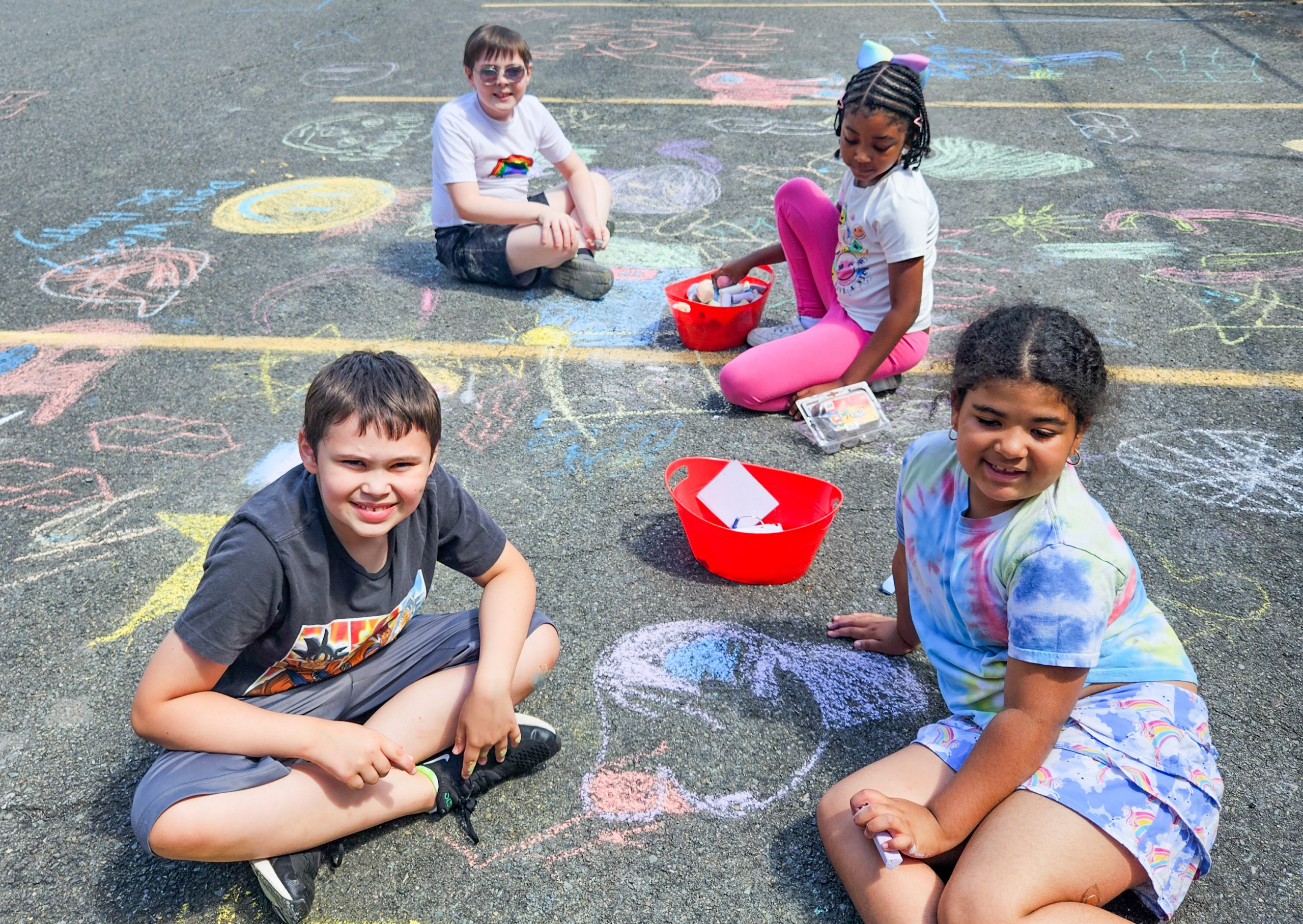 Four elementary school children sit on the pavement outside a school, playing with sidewalk chalk.